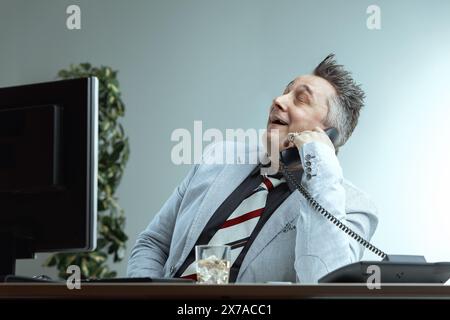 uomo di mezza età con un abito leggero e una cravatta a righe si appoggia, sorridendo con sicurezza mentre parla al telefono. La sua espressione compiaciuta e il bicchiere di whiskey Foto Stock