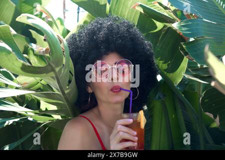 Donna tropicale nel verde denso che beve cocktail alcolici occhiali da sole. Foto Stock