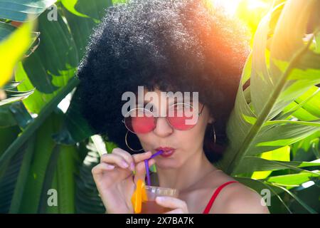 donna in una fitta vegetazione che beve cocktail alcolici con occhiali da sole. Foto Stock