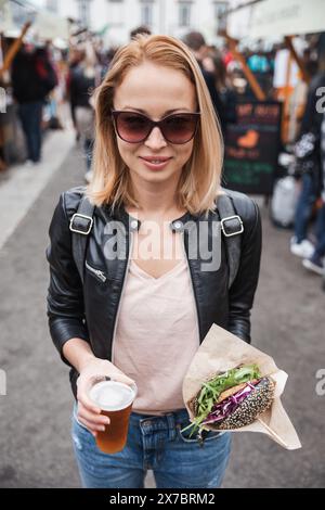 Bella giovane donna che tiene deliziosi salmone biologico vegetariano hamburger e birra IPA fatta in casa su birra all'aperto un hamburger urbano Street food festival a Lubiana, Slovenia Foto Stock