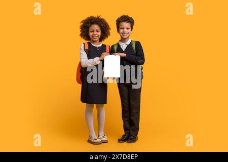 Due bambini della scuola che tengono un tablet vuoto su sfondo giallo Foto Stock