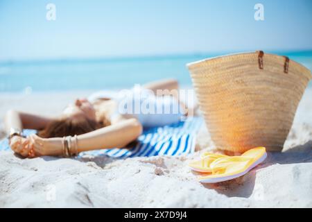 Primo piano su una moderna donna di 40 anni sulla riva dell'oceano con sacchetto di paglia, infradito e asciugamano a righe. Foto Stock