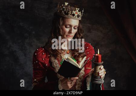 stressata regina medievale in abito rosso con libro, candela e corona su sfondo grigio scuro. Foto Stock