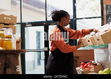 Un negoziante afroamericano che organizza un negozio ecologico con prodotti freschi, imballaggi senza plastica e prodotti sfusi. Donna nera che organizza scatole di frutta e verdura coltivate in fattoria sugli scaffali Foto Stock