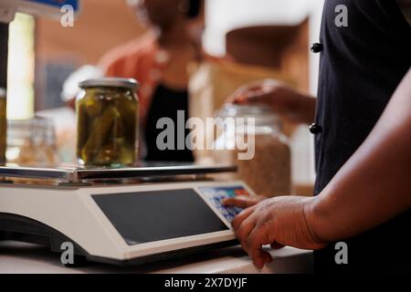 Individuo afroamericano in grembiule che assiste un cliente al check-out in un negozio di alimentari. Il barattolo di vetro di sottaceti freschi viene pesato su scala digitale dal fornitore femminile nel mercato alimentare biologico ecologico. Foto Stock