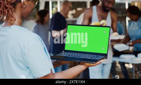 Vista dettagliata di una donna afroamericana volontaria che tiene un laptop con un display isolato per la personalizzazione. La signora nera tiene in mano il minicomputer con il modello cromakey a schermo verde. Foto Stock