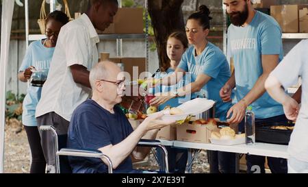 Anziano in sedia a rotelle che riceve cibo gratuito da un gruppo di volontari multietnici in occasione di un evento di soccorso contro la fame. Simpatici operatori di beneficenza che forniscono pasti caldi ai senzatetto e ai disabili. Foto Stock