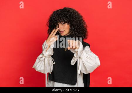 Ti sto osservando. Donna caucasica sicura e attenta con capelli ricci che puntano agli occhi e alla macchina fotografica, mostra che ti sto guardando gesto, spiando qualcuno. Ragazza severa isolata su sfondo rosso Foto Stock