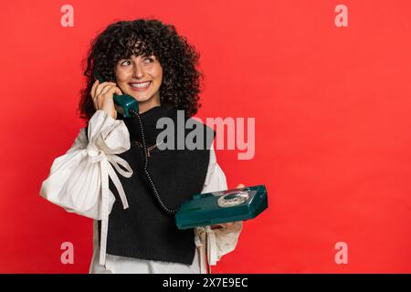 Ehi, chiamami. Giovane donna caucasica con i capelli ricci che parla su un telefono fisso fisso cablato degli anni '80, proposta pubblicitaria di conversazione, shopping online. Ragazza isolata su sfondo rosso Foto Stock