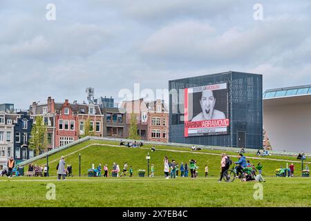 Paesi Bassi, Amsterdam - 8 aprile 2024: Le persone camminano e riposano al Museumplein, di fronte al Museo Stedelijk e al poster di Abramovic Foto Stock