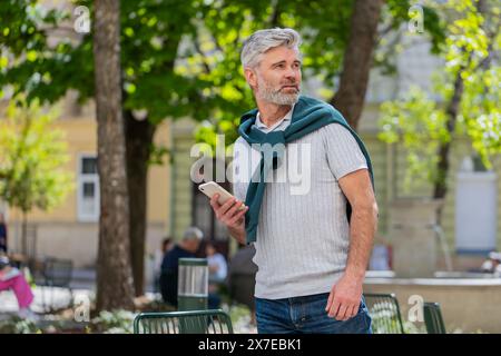 Viaggiatore turistico uomo maturo che utilizza messaggi di testo sullo smartphone in cerca di un modo di ricerca sulla mappa nell'app mobile Navigator all'aperto. Un uomo con la barba caucasica passeggia per le strade urbane della città. Foto Stock