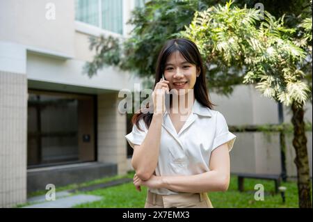 Una bella donna asiatica sorridente sta parlando al telefono con qualcuno mentre cammina in giardino o in giardino in una giornata luminosa. persone e wireless tec Foto Stock