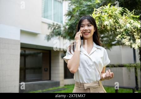 Una bella donna asiatica sorridente sta parlando al telefono con qualcuno mentre cammina in giardino o in giardino in una giornata luminosa. persone e wireless tec Foto Stock