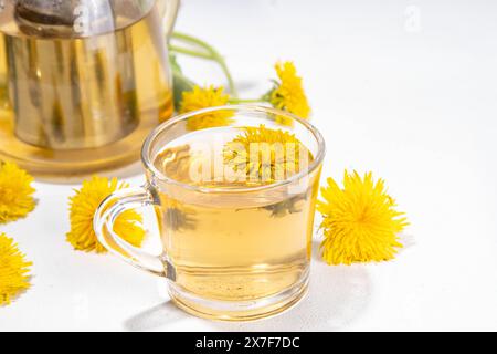 Tè di fiori a base di erbe di leoni. Tazza di vetro trasparente e teiera con bevanda dorata a base di tè caldo, su sfondo bianco con fiori e foglie di dente di leone Foto Stock