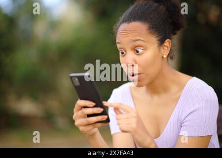 Una donna nera stupita che controlla il cellulare fuori in un parco Foto Stock