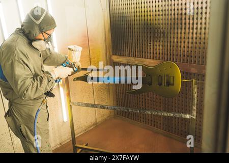 Chitarrista con uniforme protettiva in officina che utilizza la pistola a spruzzo per verniciare la chitarra, creando una nuova chitarra in fabbrica. Foto di alta qualità Foto Stock