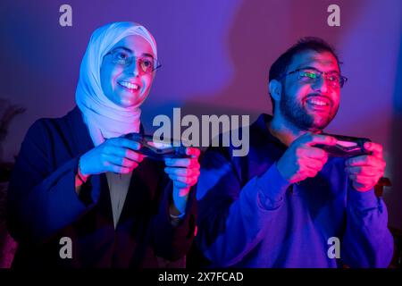 Coppia o amici hanno un viso scioccante ed emozionante con divertimento, utilizzando il joystick per gareggiare e giocare insieme, seduti in camera a casa con decorazioni al neon Foto Stock