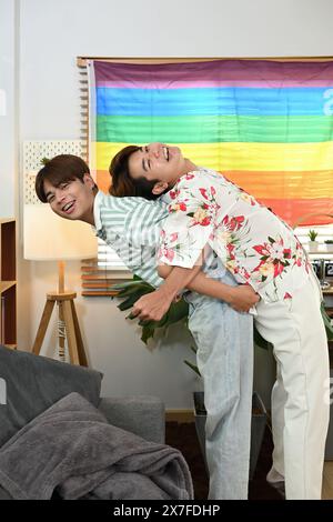 Allegra coppia gay maschio che si fanno un giro in piggyback per l'altro ridono gioiosamente in salotto Foto Stock