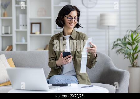 Una giovane donna asiatica con gli occhiali, sorridente mentre gestisce le bollette e le finanze utilizzando uno smartphone e un computer portatile in un salotto ben illuminato e accogliente. Foto Stock