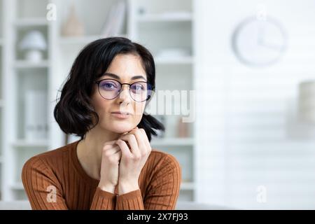 Ritratto di una donna sicura di sé con gli occhiali seduti in un elegante interno di casa, che guarda la fotocamera. Foto Stock