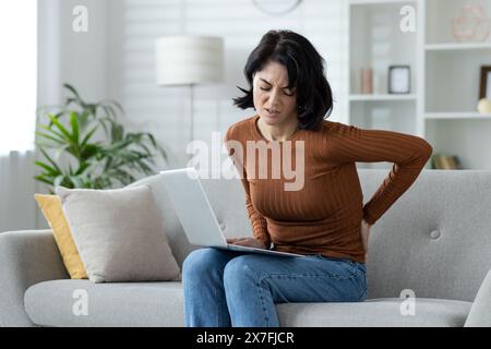 Donna che soffre di mal di schiena mentre utilizza un notebook sul divano, raffigurante disagio, postura scarsa e problemi di salute. Ideale per concetti relativi al lavoro, alla salute e all'ergonomia. Foto Stock