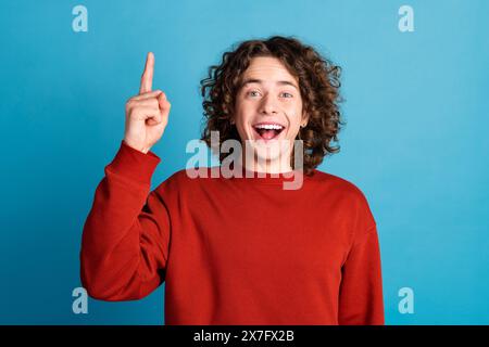 Foto di un ragazzo divertente eccitato che indossa un maglione rosso che mostra uno spazio di emtpy con le dita su sfondo blu isolato Foto Stock