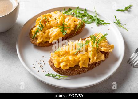 Uova strapazzate, panini e caffè per una sana colazione cheto. Tostatura integrale con uova strapazzate e rucola biologica su tavola bianca. Foto Stock