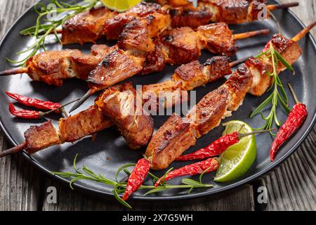 delizioso kebab di maiale succoso su piatto grigio con rosmarino, lime e peperoncino rosso secco su un tavolo rustico in legno, vista panoramica dall'alto, primo piano Foto Stock