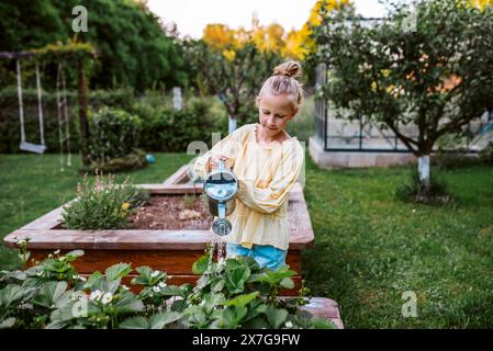 Ragazza che annaffia fragole in un letto rialzato e tiene una lattina di metallo. Cura del giardino e piantare fiori primaverili. Foto Stock