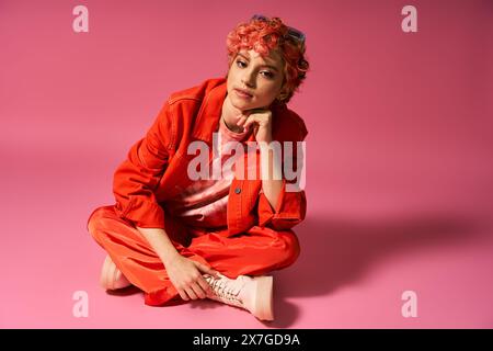 Una donna con i capelli rossi in un abbigliamento vivace si siede elegantemente su uno sfondo rosa. Foto Stock