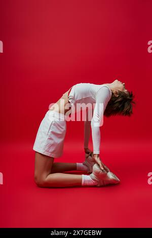 Elegante giovane donna con camicia bianca e pantaloncini che praticano yoga su uno sfondo rosso vivace. Foto Stock