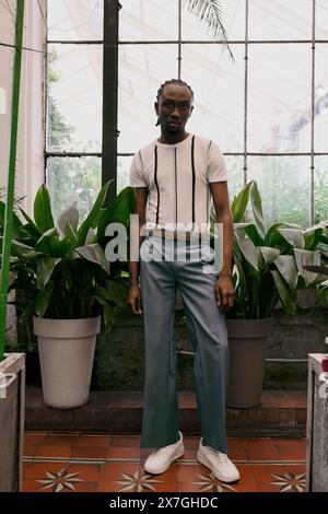 Un sofisticato uomo afro-americano in stile dapper si trova accanto a una finestra in un luminoso giardino verde. Foto Stock