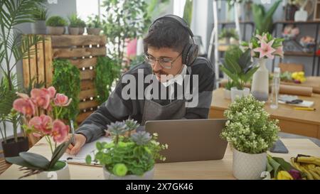 Un giovane ispanico con i baffi prende appunti in un negozio di fiori, circondato da piante vivaci e che lavora su un computer portatile. Foto Stock