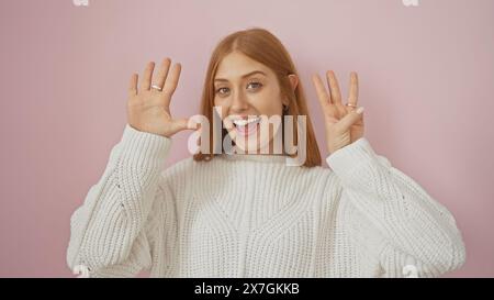 Una allegra donna caucasica con un maglione bianco fa il numero otto con le dita su uno sfondo rosa. Foto Stock