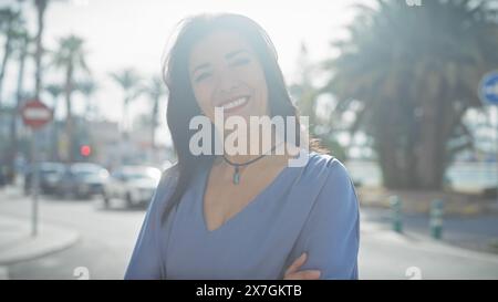 Donna sorridente in piedi con sicurezza su una strada urbana soleggiata con palme sullo sfondo. Foto Stock