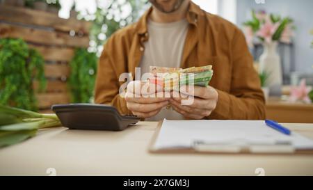 Il giovane ispanico conta le banconote in euro in un negozio di fiori, circondato da piante verdi e attrezzature da fiorista. Foto Stock