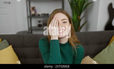 Una giovane donna caucasica sorridente con un maglione verde copre giocosamente un occhio in un soggiorno accogliente e moderno. Foto Stock