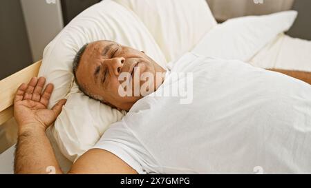 Un uomo ispanico di mezza età riposa tranquillamente in una luminosa camera da letto, emblema del relax e del comfort domestico. Foto Stock