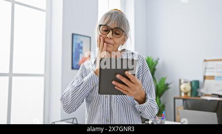 Una donna matura sorpresa con i capelli grigi legge un tablet in un luminoso interno dell'ufficio. Foto Stock