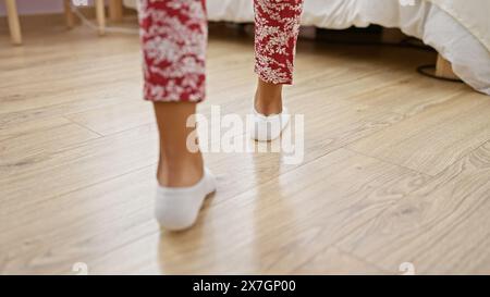 Una donna in pigiama floreale e calzini bianchi cammina attraverso una camera da letto con pavimento in legno, il che implica una routine mattutina accogliente. Foto Stock