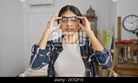 Giovane donna indiana che indossa occhiali di sicurezza in un laboratorio di falegnameria, incarna bellezza e professionalità all'interno. Foto Stock