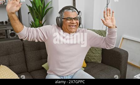 Un uomo felice di mezza età dai capelli grigi ascolta la musica e balla gioiosamente nel suo salotto di casa. Foto Stock