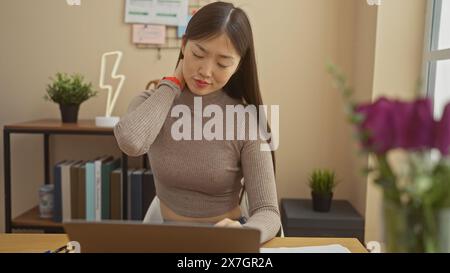 Una giovane donna asiatica soffre di dolore al collo mentre lavora sul suo notebook in un moderno ambiente domestico Foto Stock