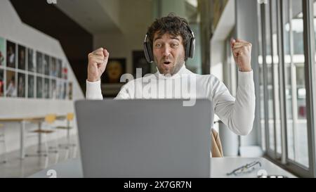 Il giovane ispanico di un ufficio reagisce con gioia davanti a un notebook indossando le cuffie Foto Stock