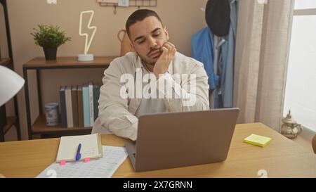 Un giovane ispanico pensivo lavora da casa su un laptop in un soggiorno ben organizzato. Foto Stock