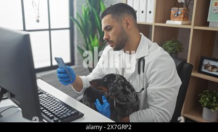 Veterinario ispanico maschile che usa un telefono cellulare mentre tiene un cane teckel in un ambiente clinico. Foto Stock
