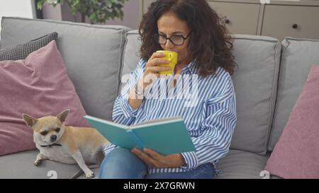 Una donna ispanica di mezza età ama leggere e bere un caffè con il suo chihuahua domestico in un accogliente soggiorno. Foto Stock