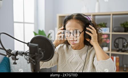 Una donna ispanica sorridente e matura regola le cuffie in un moderno studio radio con microfono e filtro pop. Foto Stock