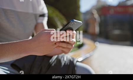 Primo piano di un giovane ispanico che tiene in mano uno smartphone in un ambiente urbano all'aperto, concentrato e con messaggi di testo. Foto Stock