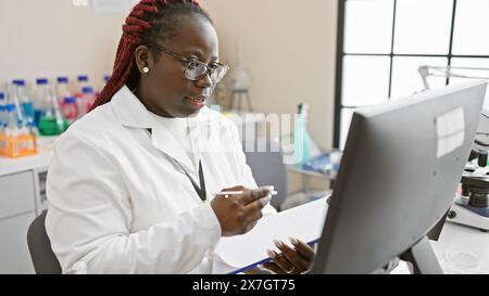 Donna afroamericana con trecce che lavora in un laboratorio, indossa un camice da laboratorio e occhiali. Foto Stock